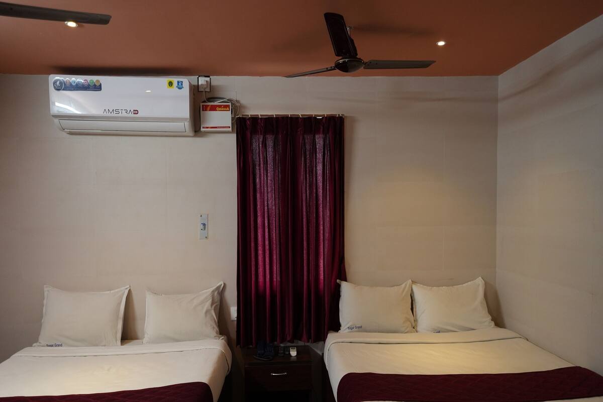 Quarto de hotel com duas camas, um ventilador de teto, aparelho de ar condicionado e janela com cortinas vermelhas.