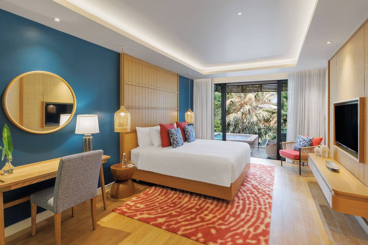 Ein modernes Hotelzimmer mit einem großen Bett, einem Schreibtisch, einem Sessel, einem Fernseher und Blick ins Freie.