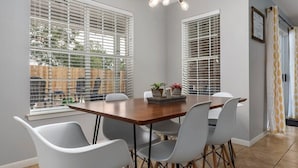 A dining area with a wooden table and six chairs, large windows with blinds, and a ceiling light fixture.
