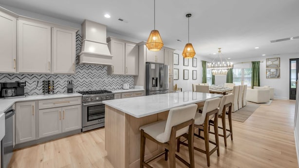 Una cocina moderna con armarios blancos, una isla central, iluminación colgante y un comedor con una mesa y sillas blancas.