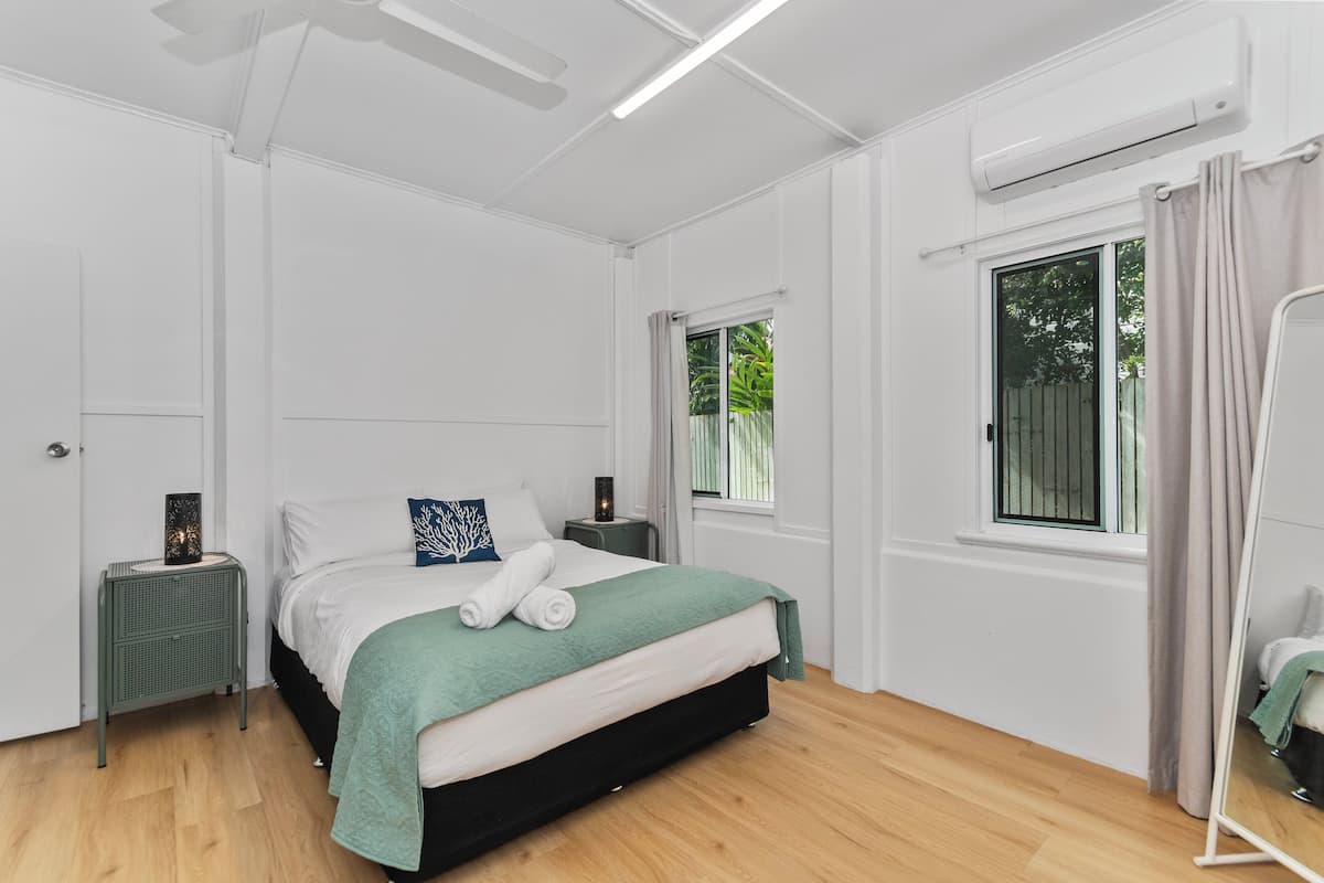 Een slaapkamer met een bed, nachtkastje, airconditioning, raam met gordijnen en een spiegel.