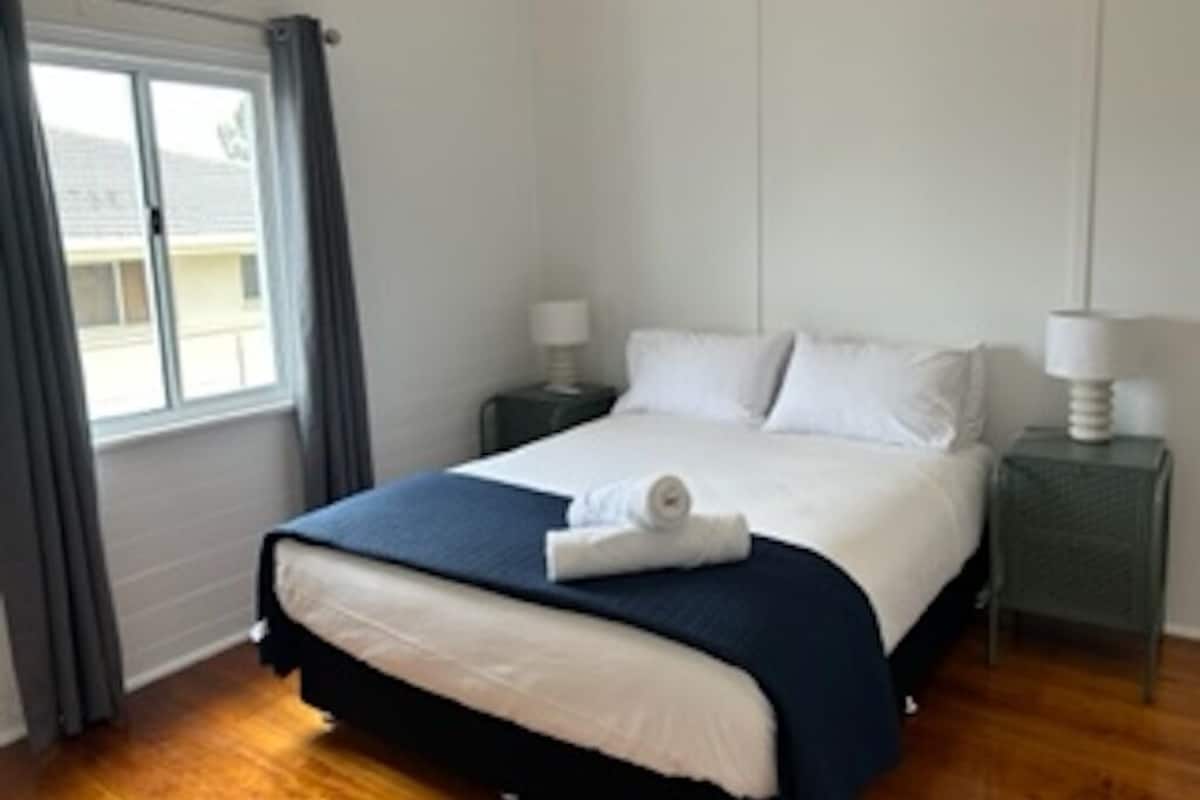 Een keurig opgemaakt bed met een blauw-witte dekbedovertrek, twee bedlampen en een raam met gordijnen.