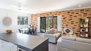 A modern kitchen with a central island, a dining area with a table and chairs, a seating area with a sofa and armchairs, and a brick-patterned wall. - Tree Views - Oceanstays (Yamba)