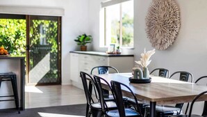 A modern dining area with a wooden table, black chairs, a hanging light fixture, and a view of greenery through a glass door. - Tree Views - Oceanstays (Yamba)