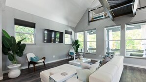 A spacious living room with a large white sofa, a coffee table, and a TV mounted on the wall. There are several windows allowing natural light to enter, and a ceiling fan is visible. - Secret Pool Cabana- Mins to Downtown and Beach (St. Augustine)