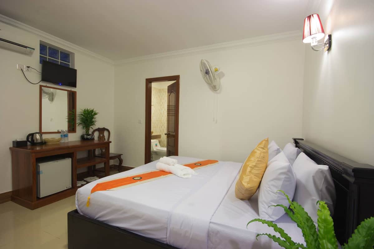Quarto de hotel com cama, escrivaninha com computador, televisão na parede, espelho, ventilador e janela.