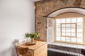 Et værelse med en stenvæg, et træbord med en vævet dækserviet og en vase med blomster, et vindue med udsigt til både, og en radiator under vinduet.