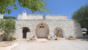 A stone building with three arched doorways and a central door, surrounded by gravel and potted plants. - Trullo Agapanthus (Ostuni)