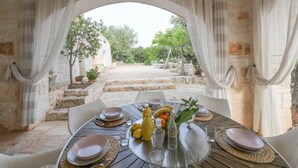 An outdoor dining area with a stone table and chairs, a view through an archway, and a table set with plates, glasses, and fruits. - Trullo Agapanthus (Ostuni)