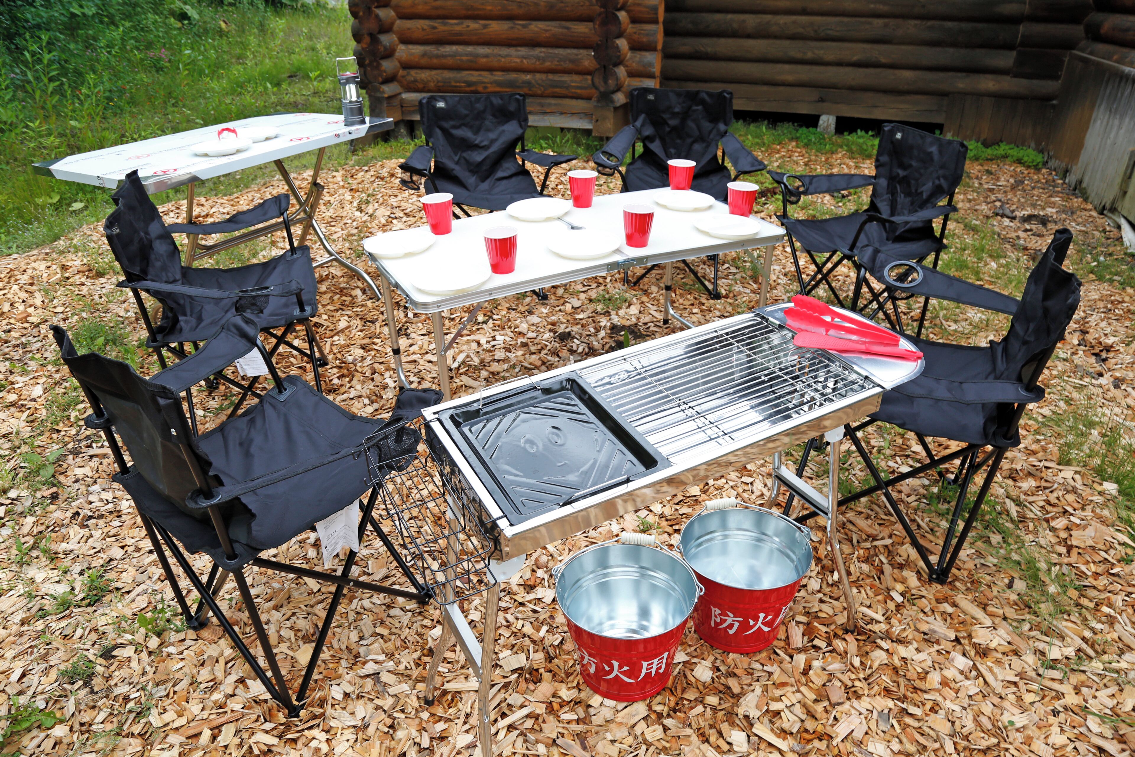 Un espace barbecue extérieur comprenant un gril, des chaises pliantes, une table et des gobelets rouges.
