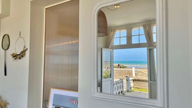 Une pièce dotée d’une grande fenêtre donnant sur une plage, d’un tableau encadré représentant une scène militaire et d’un miroir avec un cadre décoratif.