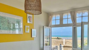 Une pièce avec un mur jaune, un canapé marron, un luminaire suspendu et une vue sur la plage par des portes ouvertes.