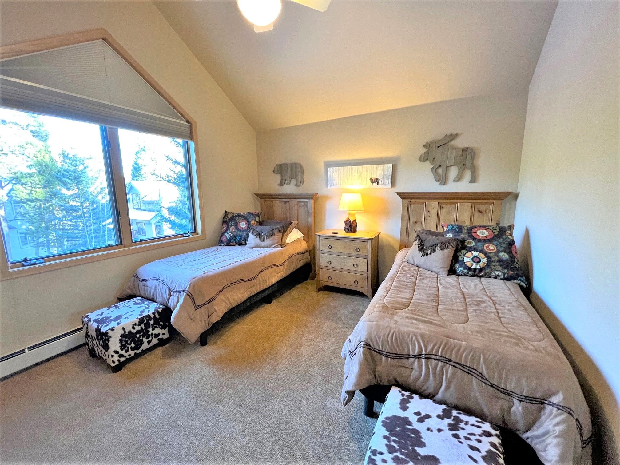 A bedroom with two beds, a nightstand with a lamp, and a window with a view of trees and houses.