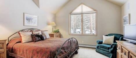Un dormitorio con techo inclinado, cama con colcha estampada, ventana con vistas a árboles, un sillón verde y un aparador de madera.