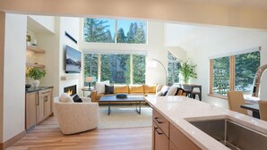 A modern kitchen with a large island, a dining area with a sofa and table, and large windows offering a view of trees and a building.