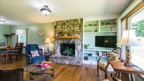 A cozy living room with a stone fireplace, wooden flooring, and a television on a shelf. - Charming 2BR w/ Fire Pit + Mountain Views + Free Tickets (Hendersonville)