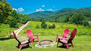 A scenic outdoor area with a fire pit and two red Adirondack chairs facing a road and green hills. - Charming 2BR w/ Fire Pit + Mountain Views + Free Tickets (Hendersonville)