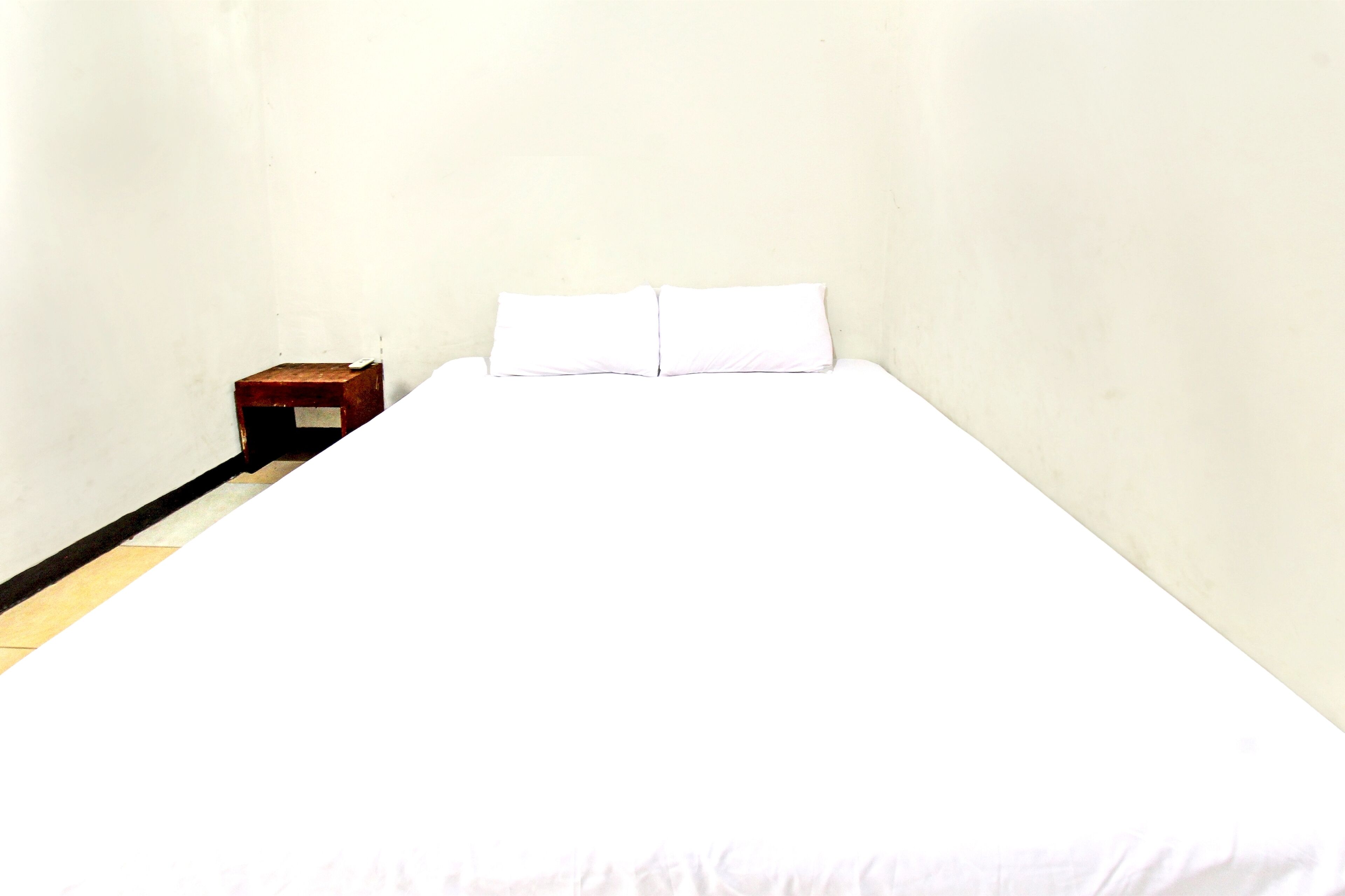 A simple, unadorned room with a bed, a small wooden nightstand, and a plain wall.