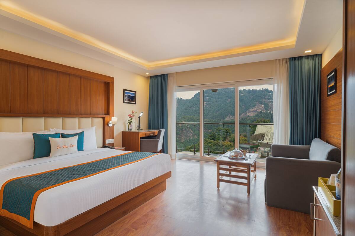 A hotel room with a large bed, a desk, a chair, and a balcony with a mountain view.