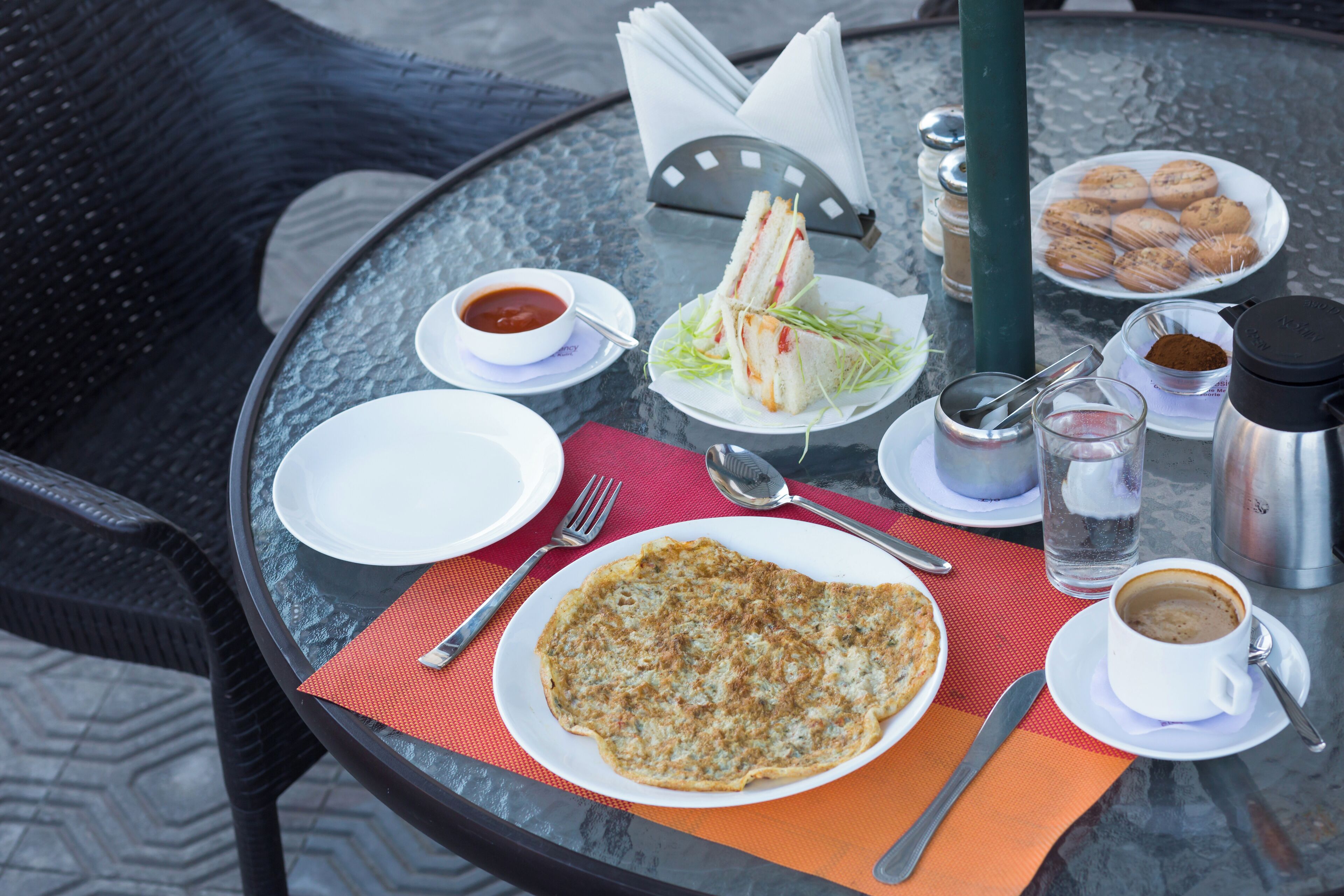 A round glass table with a variety of breakfast items including a plate of sandwiches, a plate of cookies, a bowl of sauce, a cup of coffee, and a glass of water.