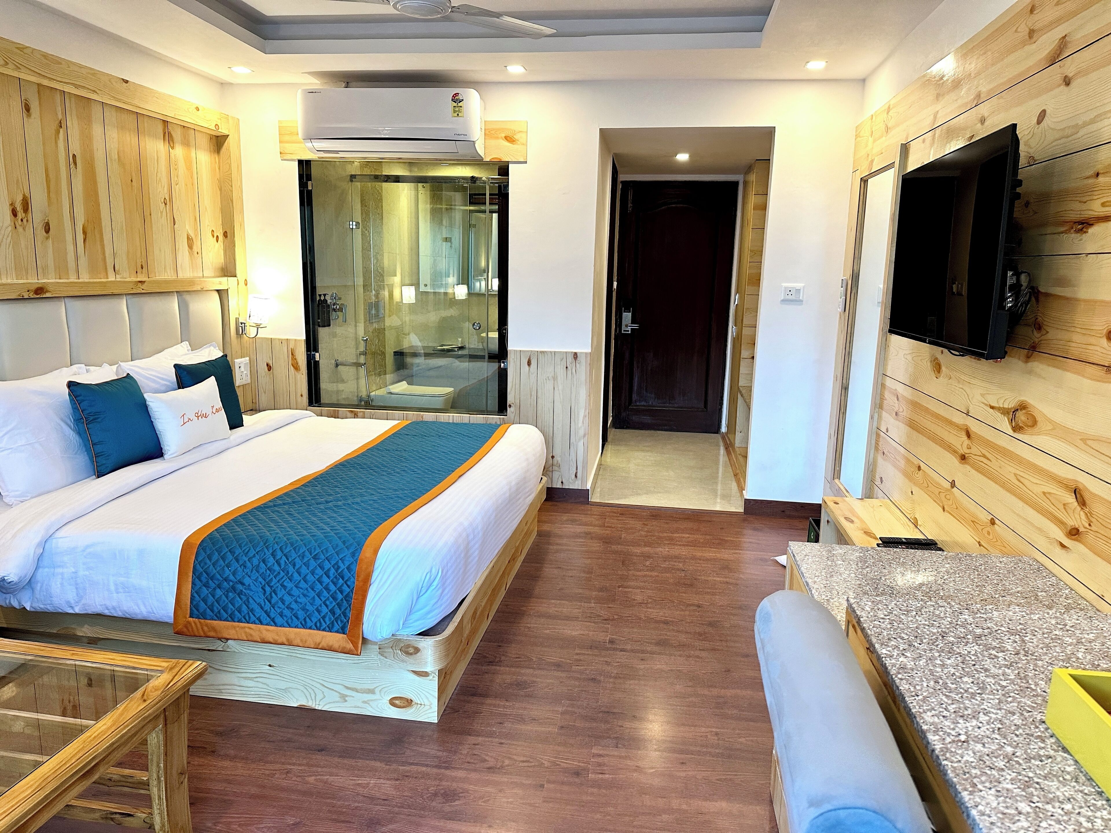 Une chambre d’hôtel avec un grand lit, une tête de lit en bois, un téléviseur fixé au mur et une salle de bain visible à travers une porte en verre.