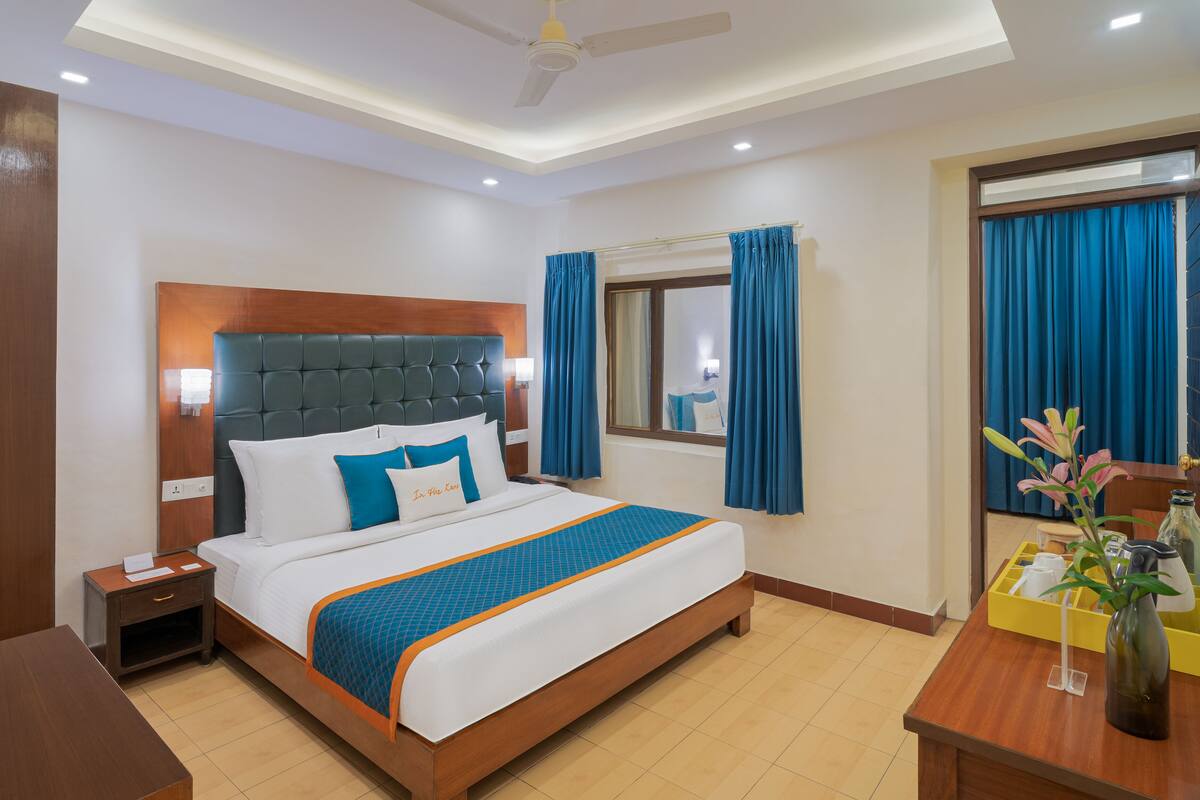 A hotel room with a large bed, wooden headboard, blue curtains, a bedside table with a lamp, and a small table with a vase of flowers.