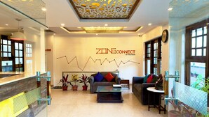 A modern reception area with a glass partition, a reception desk, a sofa with cushions, potted plants, and a wall-mounted clock. - Zone Connect by The Park Mussoorie (Dehradun)