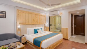 A modern hotel room with a wooden headboard, a bed with a blue and orange bedspread, a gray sofa, a small wooden table with a fruit basket, and a glass shower enclosure. - Zone Connect by The Park Mussoorie (Dehradun)