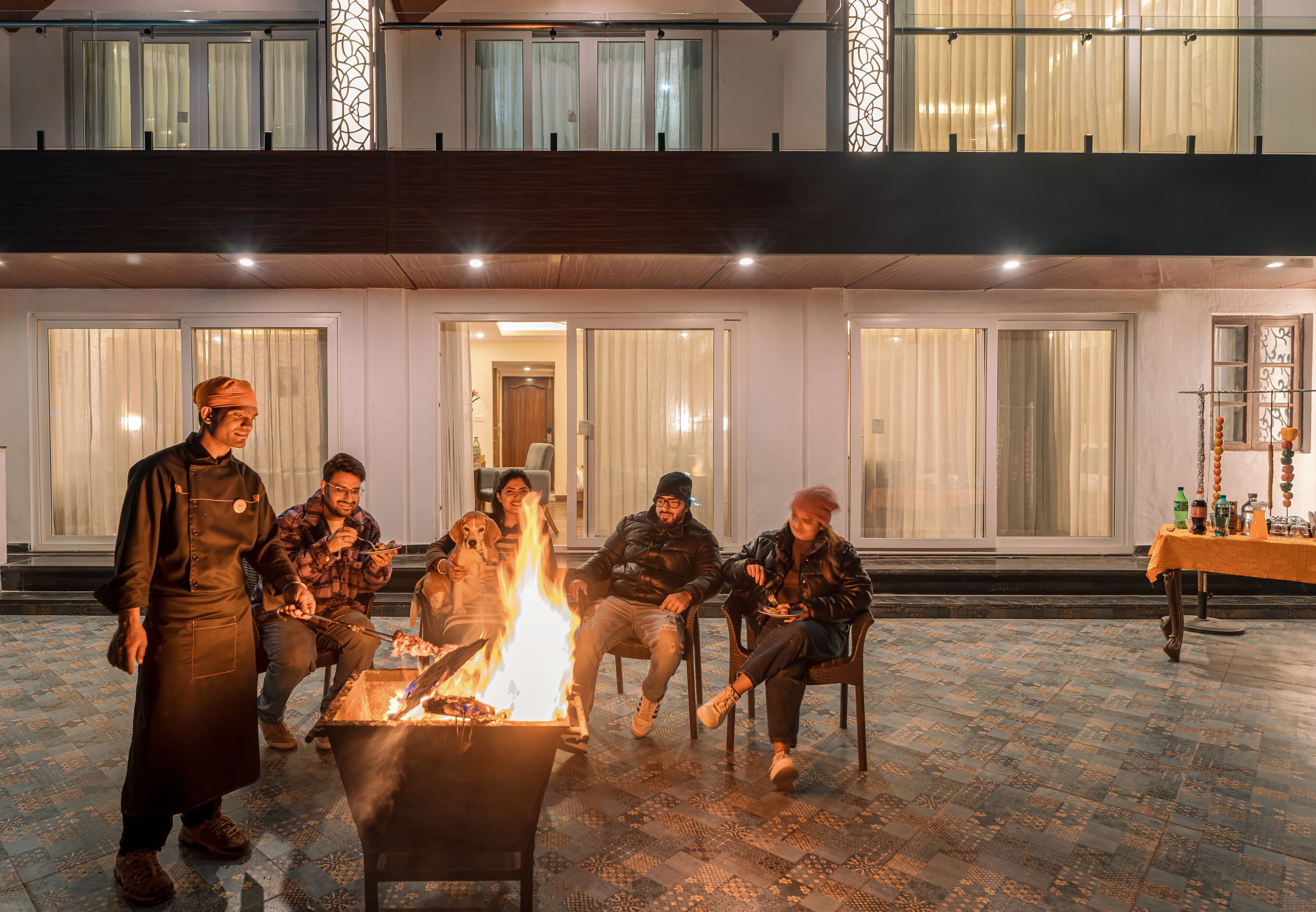 A group of people around a fire pit in an outdoor setting with a balcony and large windows.