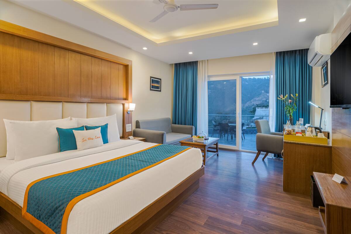 A hotel room with a large bed, a desk, a chair, a balcony with a view, and a ceiling fan.