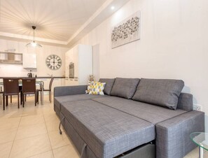 Una sala de estar moderna con un sofá gris, una zona de comedor con una mesa y sillas de vidrio, y una cocina al fondo.