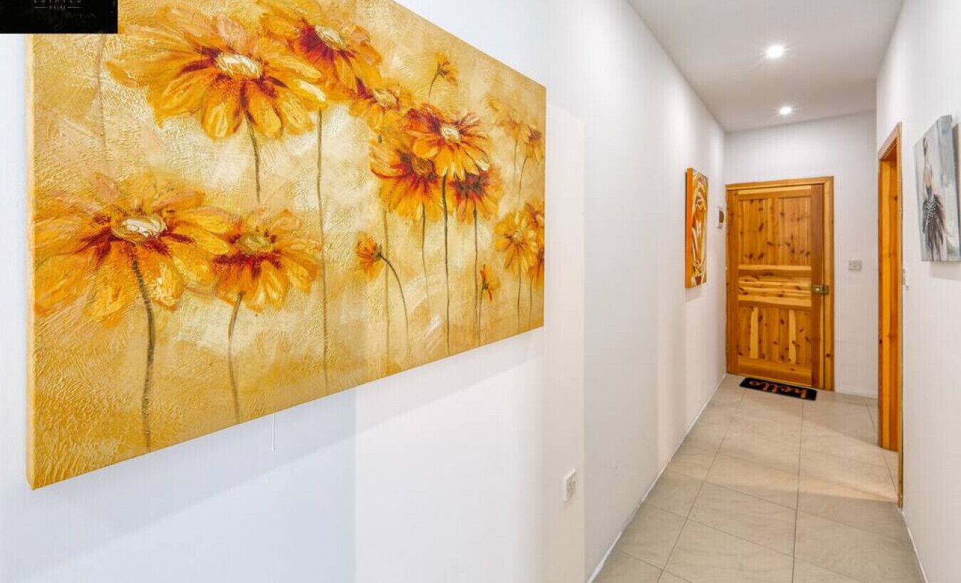 Un corridoio con un grande quadro floreale sulla parete sinistra, una porta in legno in fondo e un piccolo tappeto sul pavimento.