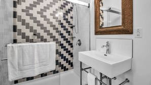A modern bathroom with a tiled shower wall, a white sink, and a large mirror with an ornate frame. - Webber Hotel (Lyndonville)