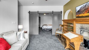 A modern hotel room with a grey sofa, a wooden side table, a fireplace, and a television. - Webber Hotel (Lyndonville)