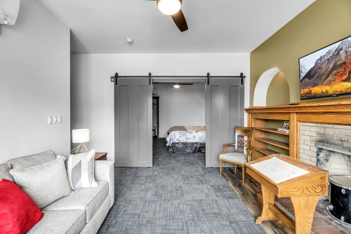A modern hotel room with a grey sofa, a wooden side table, a fireplace, and a television.