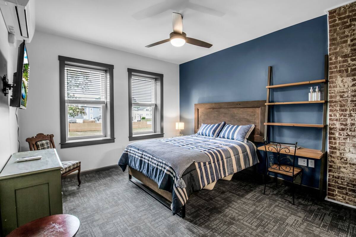 A bedroom with a bed, two windows, a ceiling fan, a desk, a chair, and a brick wall.