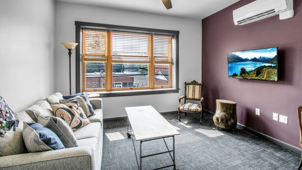 A modern living room with a sofa, a coffee table, a TV mounted on the wall, a ceiling fan, and a window with blinds. - Webber Hotel (Lyndonville)