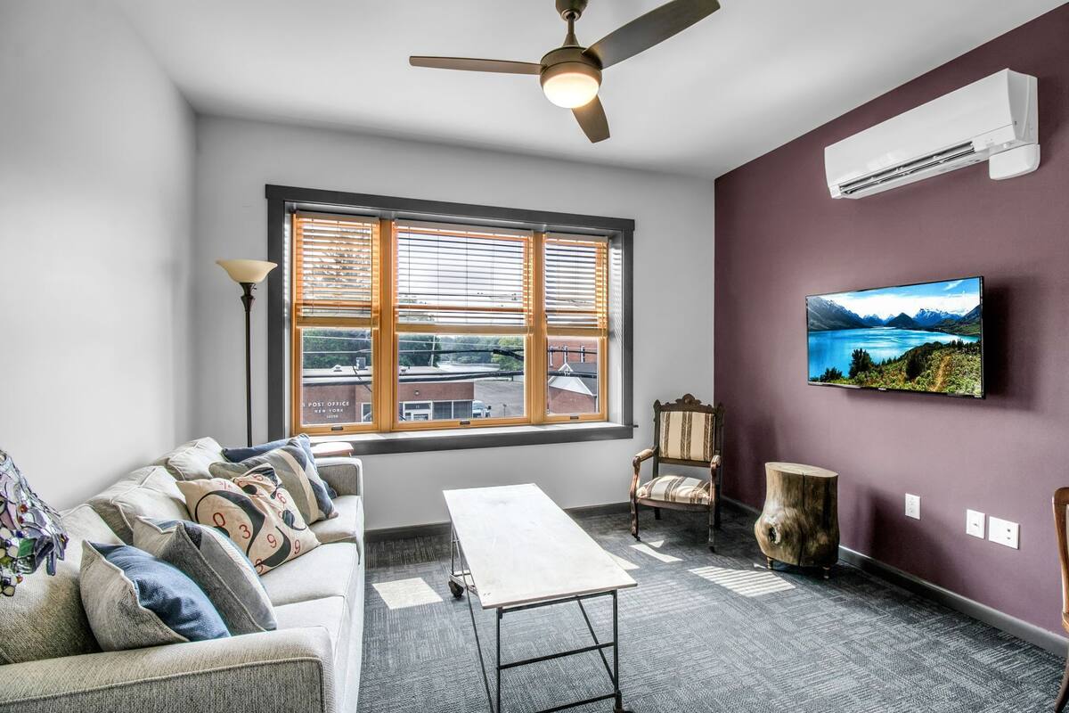 A modern living room with a sofa, a coffee table, a TV mounted on the wall, a ceiling fan, and a window with blinds.