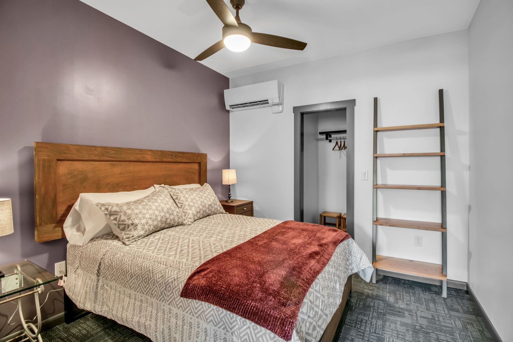 Une chambre avec une tête de lit en bois, une couvre-lit à motifs, un plaid rouge, un ventilateur de plafond, un appareil de climatisation, une échelle en bois et une petite table avec une lampe.