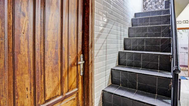 A wooden door with a metal handle, adjacent to a staircase with black tiled steps and white walls. - OYO Life 92785 Wisma Trubus  Syariah (Tuban)