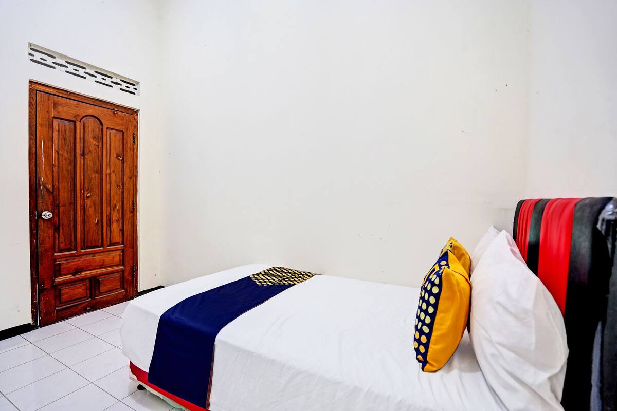 A single bed with a white mattress, a blue and red bedspread, and a yellow polka-dotted pillow. There is a wooden door with a decorative panel above it.