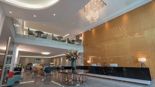 A modern hotel lobby with a glass balustrade, a central round table with a vase of flowers, a reception desk, and a mezzanine level with seating. - Stay Easy Plus Hotel (Addis Ababa)