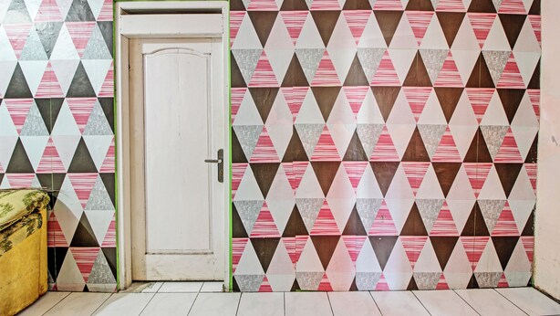 Una habitación con papel tapiz de estampado geométrico, una puerta blanca y un sillón amarillo.
