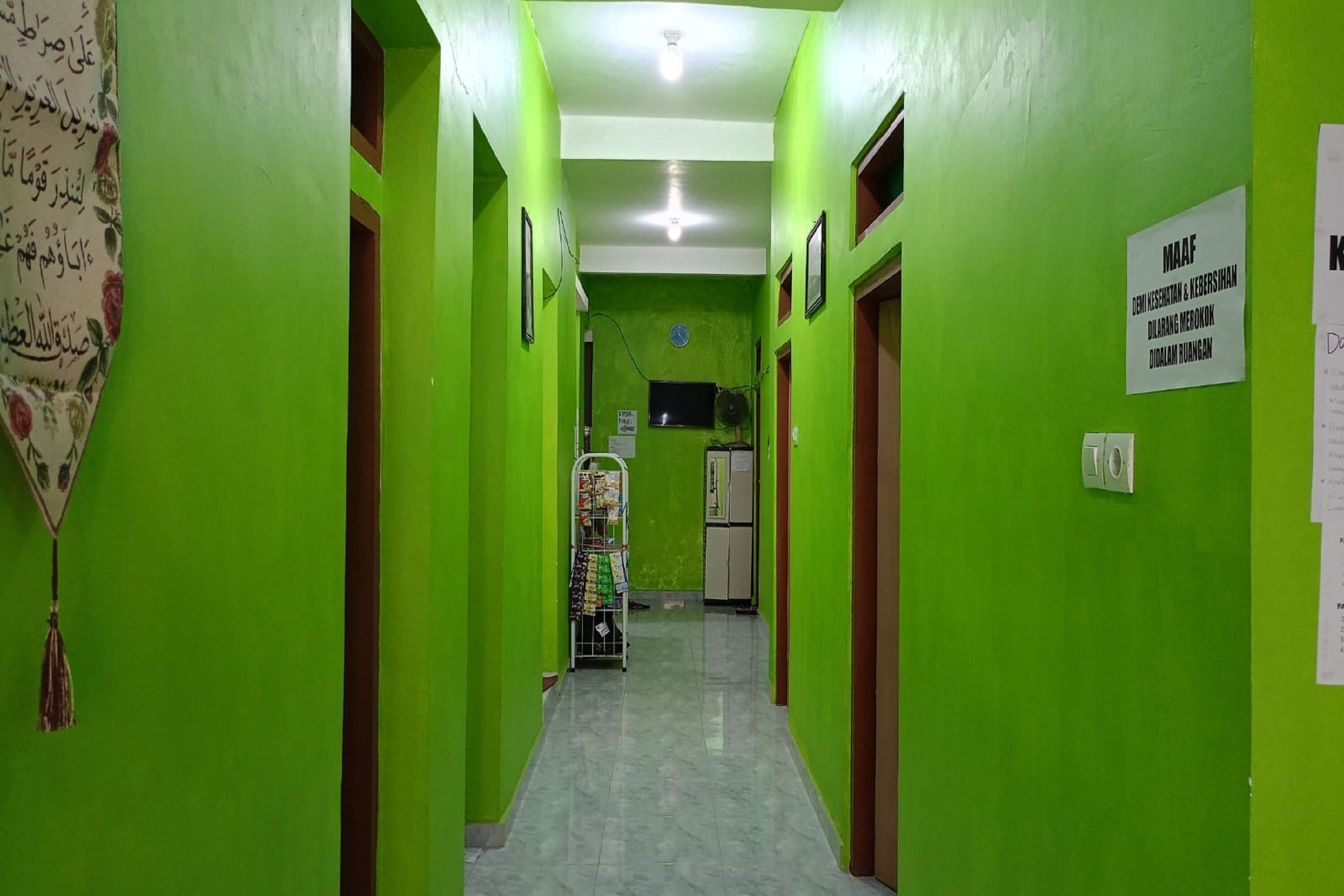 Un pasillo estrecho con paredes verdes y suelo de baldosas. Hay un cartel en la pared derecha y un ventilador en la izquierda.