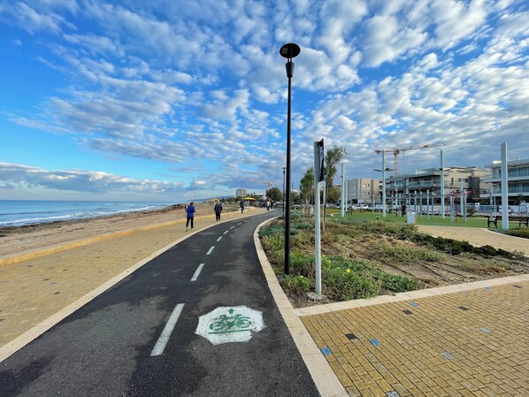 A paved path with a bike lane, a beach, and modern buildings in the background. - Breezot Yam (Nahariya)