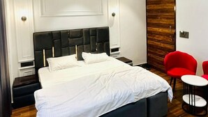 A modern bedroom with a large bed, a wooden wardrobe, a ceiling fan, and a small red chair. - Hotel Primrose - Patiala (Pantran)