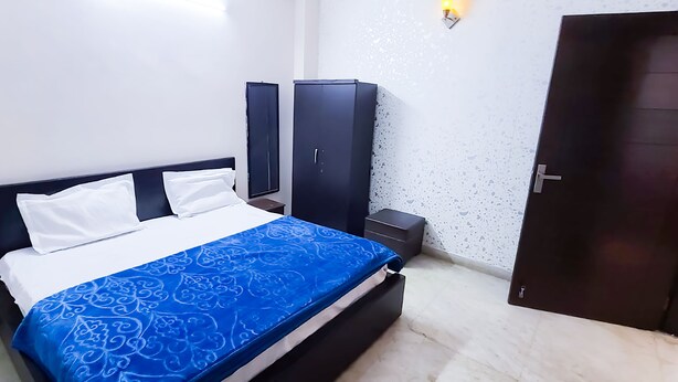 Un lit bien fait, recouvert d’une couvre-lit à motifs bleus, avec un oreiller blanc et une tête de lit sombre. Une petite table de chevet avec une lampe se trouve à côté du lit, et une armoire de couleur foncée est adossée au mur.