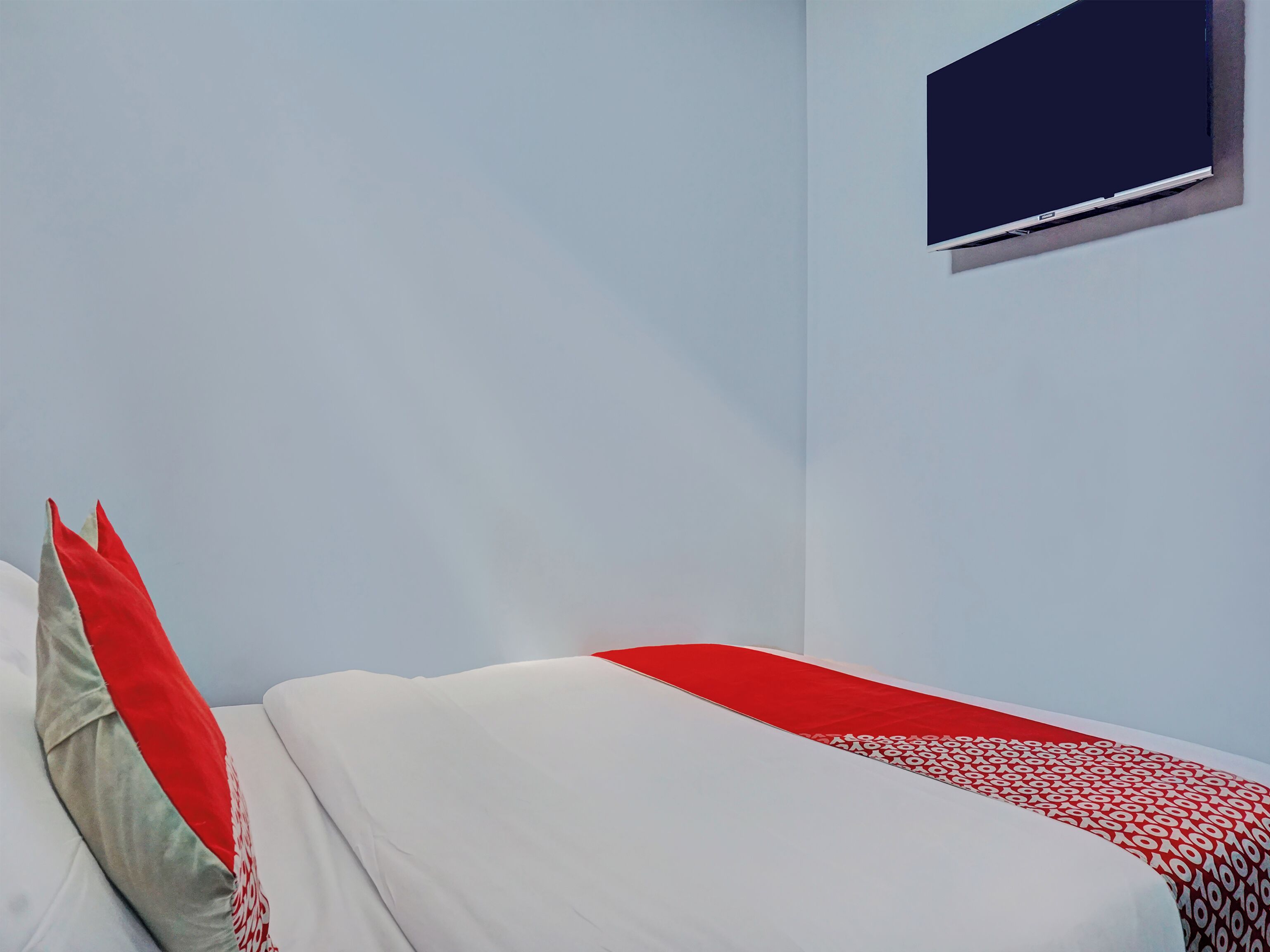 Un dormitorio con cama, almohadas, un televisor montado en la pared y una colcha de diseño rojo y blanco.