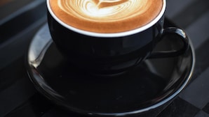 A cup of coffee with latte art on a black saucer on a reflective surface. - Roomshala 103 Good Luck House (New Delhi)