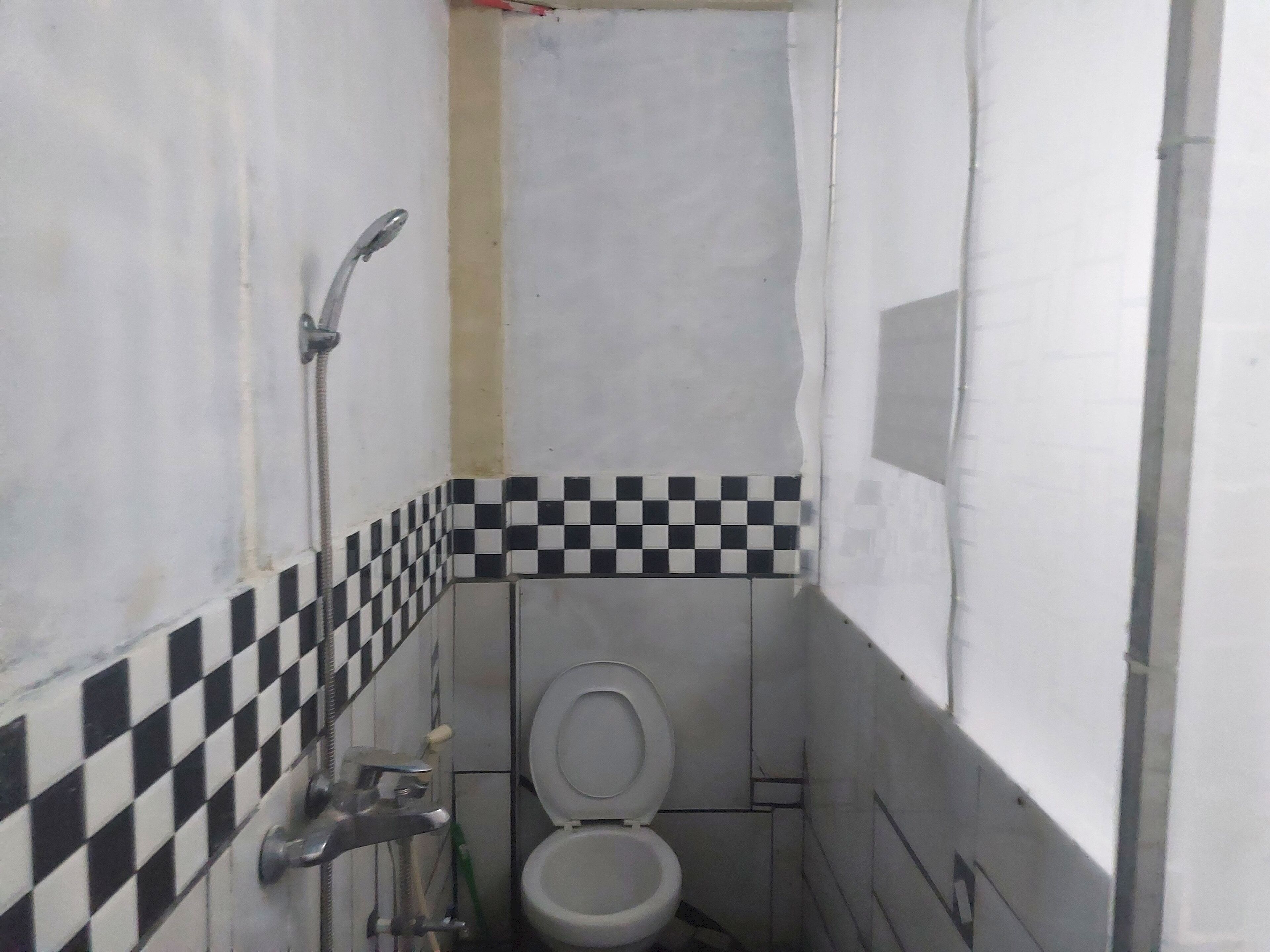 A small, narrow bathroom with a toilet, a shower, and a checkered tile border.